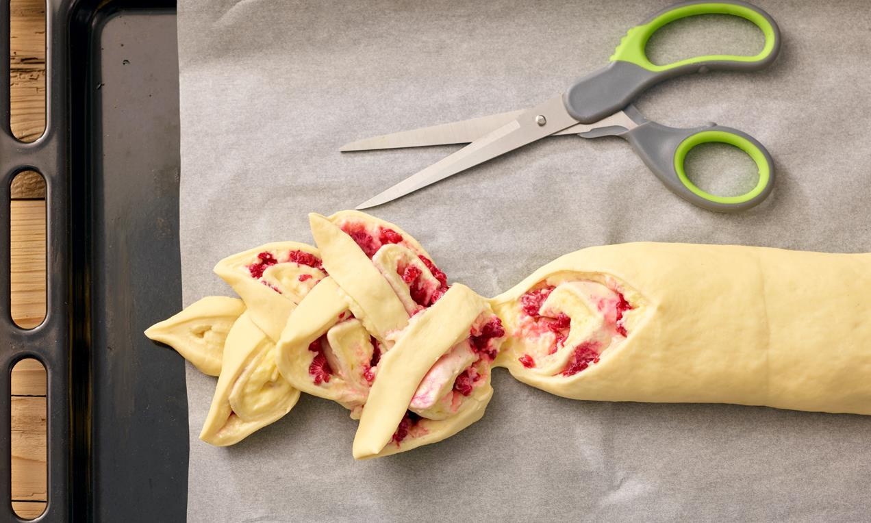 A Pudingos-málnás kalács sütemény tésztáját konyhai ollóval vágjuk szeletekre sütés előtt formázva meg belőlük a süteményt.