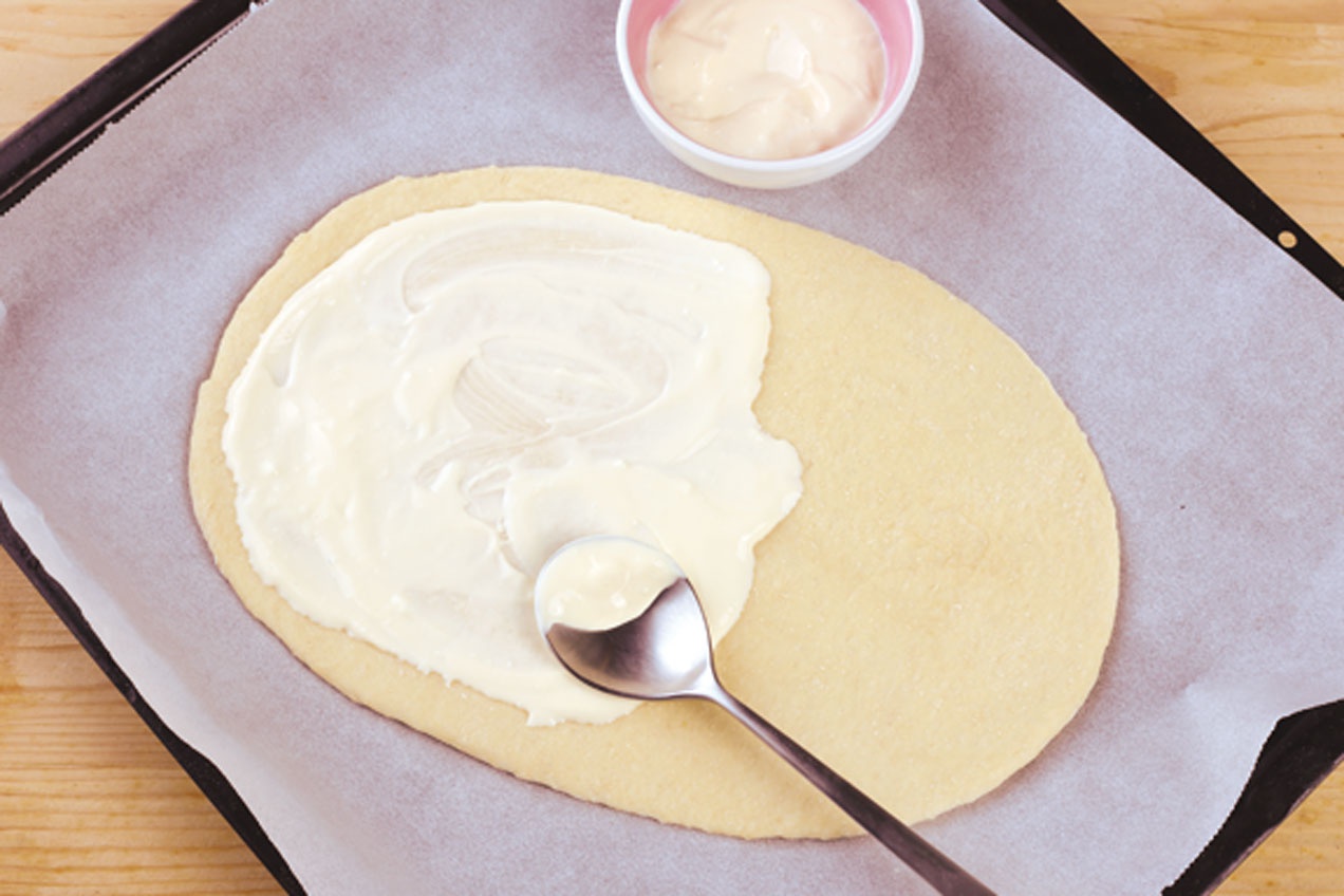 Az Édes pizza sütemény tésztáját a feltétek előtt megkenjük a tejfölkeverékkel.