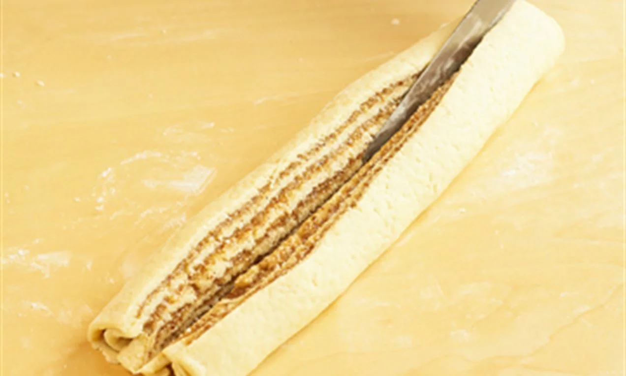 A Mákos fonat tésztáját kinyújtjuk, majd megkenjük töltelékkel és feltekerjük, majd ketté vágjuk.